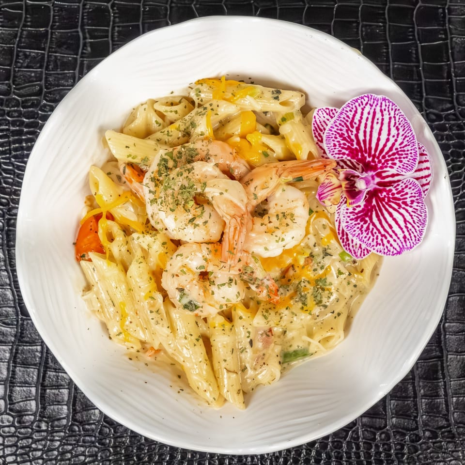 Creamy Jamaican shrimp pasta with bell peppers, herbs, and an orchid garnish in a white bowl.