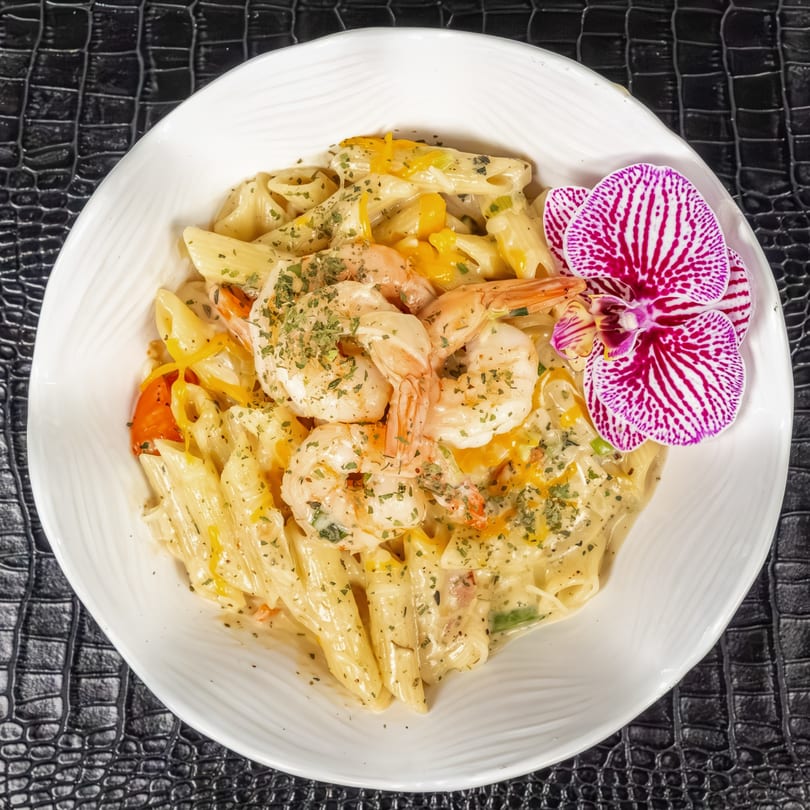 Creamy Jamaican shrimp pasta with bell peppers, herbs, and an orchid garnish in a white bowl.